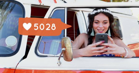 Téléchargez les photos : Gros plan d'une hippie blanche penchée sur la porte d'une camionnette alors qu'elle tapait sur son téléphone. À côté d'elle au premier plan se trouve une image numérique d'une bulle de message avec une icône cardiaque qui augmente en nombre - en image libre de droit