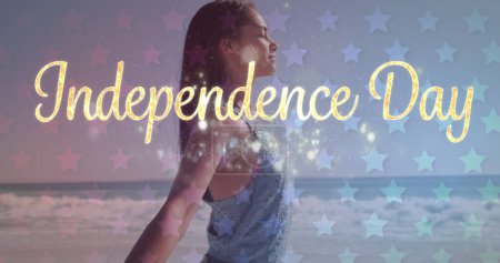 Young biracial woman celebrates Independence Day at the beach. Sparkling graphics overlay the scene, enhancing the festive mood.