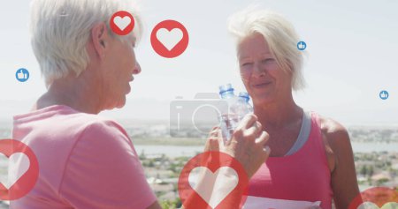 Image of social media icons, over happy senior women drinking water and laughing on beach. social media, fitness, healthy retirement and communication network concept, digitally generated image.