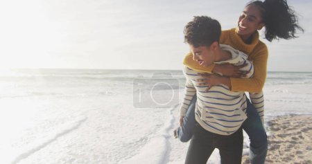 Photo for Biracial woman gives piggyback ride to young biracial man on a beach. Their joyful moment captures the essence of carefree outdoor leisure by the sea. - Royalty Free Image