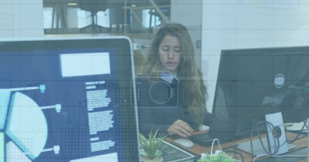 Photo for In this image, we see a biracial businesswoman working in a modern office. She is sitting at a desk, working on a computer. Her female colleague is working next to her. Financial data processing over a grid network. - Royalty Free Image