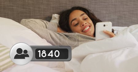 Photo for Close up of a Hispanic woman lying in bed smiling while checking her phone. Beside her in the foreground is a digital image of a friend request icon increasing in count - Royalty Free Image