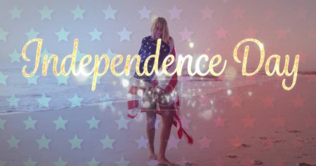 Foto de Joven mujer caucásica envuelta en una bandera americana en una playa. Sparkles y superposición de texto del Día de la Independencia significan una celebración patriótica. - Imagen libre de derechos