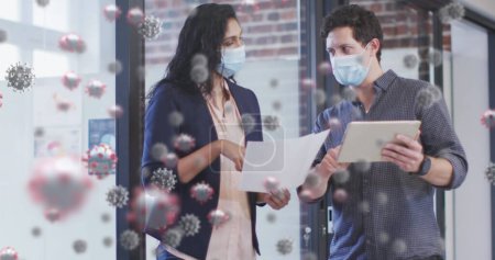 Bild von covid 19 Zellen über Kollegen in Gesichtsmasken mit digitalem Tablet im Büro. Gesundheitsfürsorge und Schutz während der Coronavirus-Covid 19 Pandemie, digital generiertes Bild.