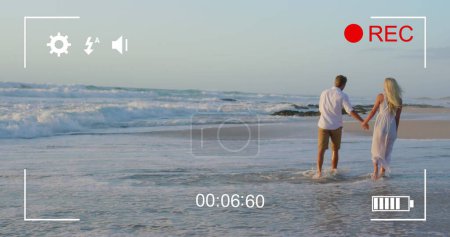 Foto de Imagen de una joven pareja caucásica caminando por una playa cogida de la mano, vista en una pantalla de una cámara digital en modo de grabación con iconos y temporizador 4k - Imagen libre de derechos