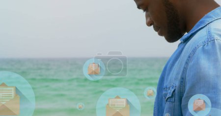 Téléchargez les photos : Image d'icônes de courrier sur le milieu de l'homme biracial à l'aide d'un smartphone à la plage. connexions mondiales, médias sociaux, technologie et concept d'interface numérique image générée numériquement. - en image libre de droit