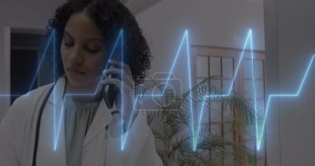 Image of cardiograph over happy biracial female doctor talking on smartphone. Medicine, health and digital interface concept, digitally generated image.