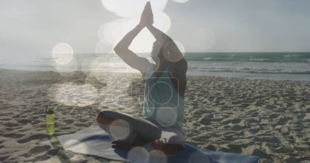Téléchargez les photos : Image de lumières brillantes sur un homme afro-américain faisant de la méditation de yoga sur la plage. Connexions mondiales, bien-être, forme physique et mode de vie sain concept image générée numériquement. - en image libre de droit