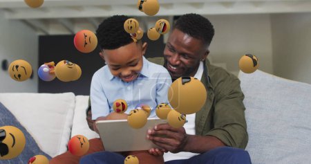 Bild von Emoticons über glückliche afrikanisch-amerikanische Vater und Sohn mit Tablet auf dem Sofa. Familie, Zweisamkeit, qualitativ hochwertiges Zeitkonzept digital generiertes Bild.