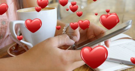 Photo for Close up of a pair of female hands texting beside a cup of coffee and a pen and notebook. Digital hearts are flying in the foreground 4k - Royalty Free Image