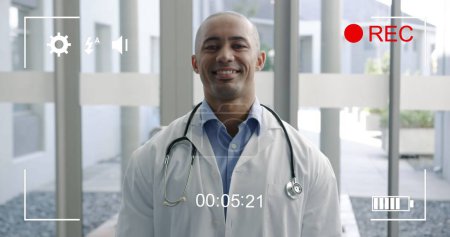 Téléchargez les photos : Image d'un portrait d'un jeune médecin masculin biracial souriant à la caméra, vue sur un écran d'un appareil photo numérique en mode enregistrement avec icônes et minuterie - en image libre de droit