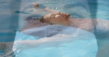 Photo for Image of Asian woman wearing a face mask, looking at the camera over Caucasian woman swimming. Coronavirus Covid-19 pandemic concept digital composite - Royalty Free Image