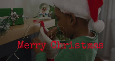Image of merry christmas text over african american boy wearing santa hat. Christmas, tradition and celebration concept digitally generated image.