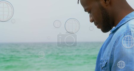 Téléchargez les photos : Image de globes au-dessus du milieu de l'homme afro-américain utilisant un smartphone à la plage. connexions mondiales, médias sociaux, technologie et concept d'interface numérique image générée numériquement. - en image libre de droit