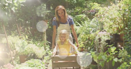 Téléchargez les photos : Spots de lumière contre la mère caucasienne portant sa fille dans le jardin cartin le jardin. semaine jardin communautaire concept de sensibilisation - en image libre de droit