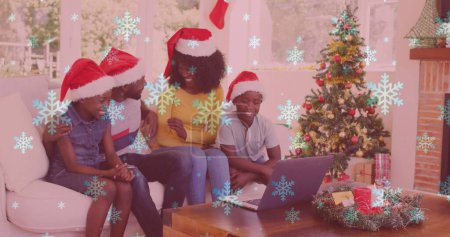 Téléchargez les photos : Image de neige tombant sur une famille souriante avec des chapeaux de Père Noël à l'aide d'un ordinateur portable. Noël, hiver, tradition et concept de célébration image générée numériquement. - en image libre de droit