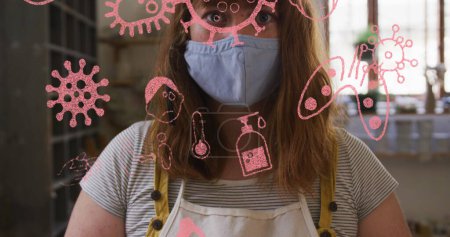 Téléchargez les photos : Image d'icônes et de cellules covid sur une femme portant un masque facial tenant de l'argile dans un atelier de poterie. global covid 19 pandémie concept image générée numériquement. - en image libre de droit