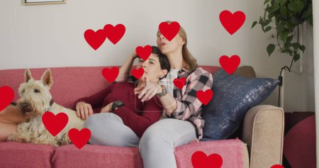 Photo for Image of hearts over caucasian female couple sitting on sofa with their pet dog. Valentines, love and celebration concept digitally generated image. - Royalty Free Image