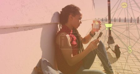 Foto de Globo de iconos digitales contra mujer afroamericana usando smartphone sentado en el paseo marítimo. concepto global de redes y tecnología - Imagen libre de derechos