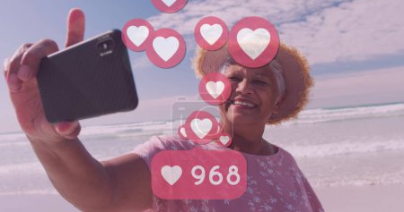 Photo for Image of hearts over happy senior african american woman taking selfie on sunny beach. healthy and active beach holiday concept digitally generated image. - Royalty Free Image