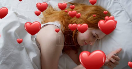 Foto de Compuesto digital de una mujer de pelo rojo caucásico acostada en la cama mientras envía mensajes de texto y corazones digitales volando en primer plano 4k - Imagen libre de derechos