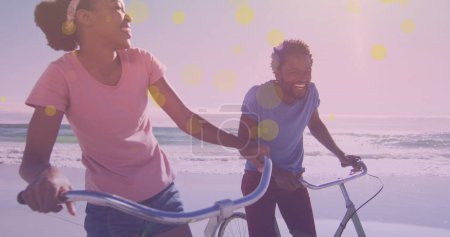 Foto de Manchas amarillas de luz contra una pareja afroamericana sentada en sus bicicletas sonriendo en la playa. concepto de conciencia del día del pedal - Imagen libre de derechos
