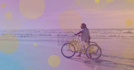 Téléchargez les photos : Des taches jaunes de lumière contre un couple de personnes âgées afro-américaines avec des vélos marchant ensemble sur la plage. concept de sensibilisation à la journée pédale - en image libre de droit