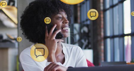 Foto de Múltiples iconos de mensajes flotando contra la mujer afroamericana hablando en el teléfono inteligente en la oficina. concepto global de redes y tecnología empresarial - Imagen libre de derechos