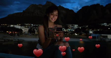 Téléchargez les photos : Composite numérique d'une femme caucasienne assise sur un bord près de la plage la nuit et les cœurs numériques au premier plan - en image libre de droit