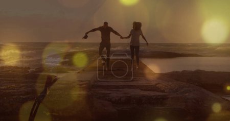 Téléchargez les photos : Taches jaunes de lumière sur le couple afro-américain tenant la main marchant sur les rochers près de la mer. Concept d'amour et de relation - en image libre de droit