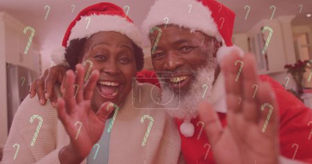 Image of candy canes falling over smiling couple with santa hats waving hands. christmas, winter, tradition and celebration concept digitally generated image.