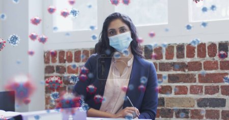 Photo for Image of covid 19 cells over woman wearing face mask, sitting by desk in office. Healthcare and protection during coronavirus covid 19 pandemic, digitally generated image. - Royalty Free Image