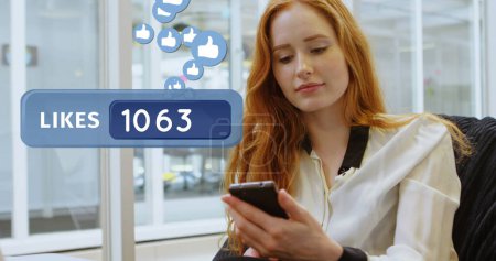 Photo for Close up of a red haired Caucasian woman texting in the office. Beside her in the foreground is a likes count bar with like icons flying upwards - Royalty Free Image