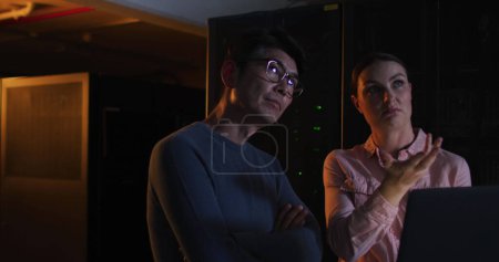 Foto de Mujer caucásica y hombre asiático revisan los datos en una sala de servidores tenue. Sus expresiones enfocadas sugieren un análisis crítico de los problemas de red en la oficina. - Imagen libre de derechos