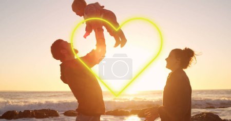 Foto de Imagen de corazón sobre padres caucásicos felices con bebé en la playa. Día internacional de las familias y concepto de celebración de imagen generada digitalmente. - Imagen libre de derechos