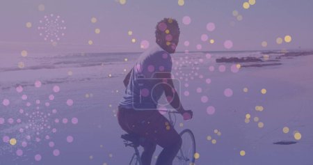 Foto de Manchas amarillas de luz contra el retrato del hombre afroamericano montando una bicicleta en la playa. concepto de conciencia del día del pedal - Imagen libre de derechos