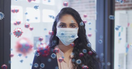 Bild von covid 19 Zellen über birassische Frau, die Gesichtsmaske im Büro aufsetzt. Gesundheitsfürsorge und Schutz während der Coronavirus-Covid 19 Pandemie, digital generiertes Bild.