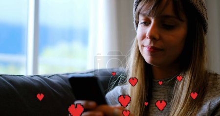 Téléchargez les photos : Composite numérique d'une femme caucasienne assise sur un canapé tout en envoyant des SMS et des cœurs numériques au premier plan - en image libre de droit