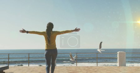 Foto de Manchas de luz contra la vista trasera de la mujer afroamericana con los brazos abiertos en el paseo marítimo. concepto de deporte y fitness - Imagen libre de derechos