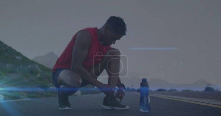 Téléchargez les photos : Image de traînées de lumière bleue contre un homme afro-américain en forme attachant des lacets de chaussures sur la route. Concept de technologie du sport et fitness, - en image libre de droit