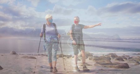 Photo for Caucasian senior couple in face masks hiking on the coast, over moving sea. active, healthy retirement and freedom during coronavirus pandemic concept,, digitally generated image. - Royalty Free Image