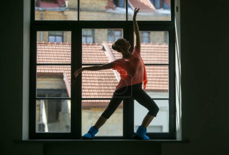 Foto de Silhouette of a young ballerina on the window sill, roof in the background. Concept of healthy lifestyle - Imagen libre de derechos