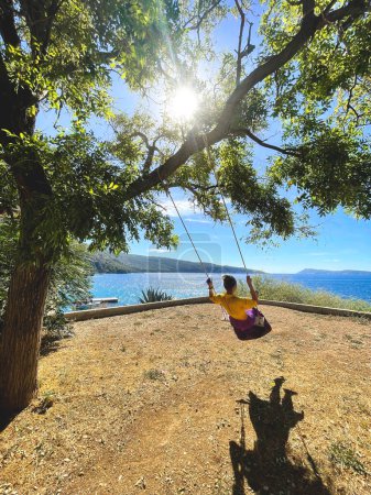 Téléchargez les photos : Happy lady rides on a swing tied to a tree behold the beautiful summer landscape of the sea and islands. - en image libre de droit