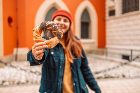 Photo for Fall tourist woman in a bright hat and autumn jacket holding baked obwarzanek traditional polish cuisine snack bagel on old city Market square in Krakow - Royalty Free Image