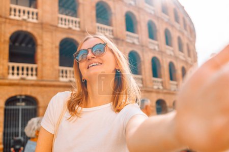 Téléchargez les photos : Jeune fille touriste en lunettes de soleil faisant selfie photo sur son smartphone en face de la célèbre Plaza de Toros de Valencia. Voyage à Barcelone concept. - en image libre de droit