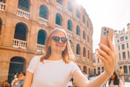 Téléchargez les photos : Jeune fille touriste en lunettes de soleil faisant selfie photo sur son smartphone en face de la célèbre Plaza de Toros de Valencia. Voyage à Barcelone Espagne concept. - en image libre de droit