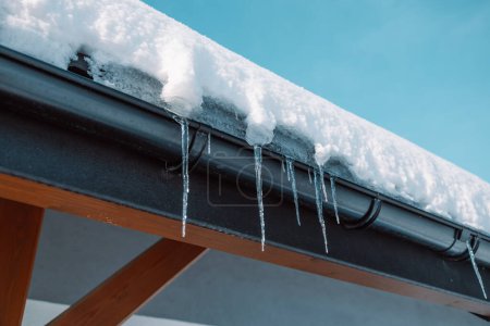 Téléchargez les photos : Des glaçons sont suspendus au toit d'une maison en rondins près de laquelle une voiture est garée négligemment, ce qui peut être endommagé par la chute de floes de glace. - en image libre de droit
