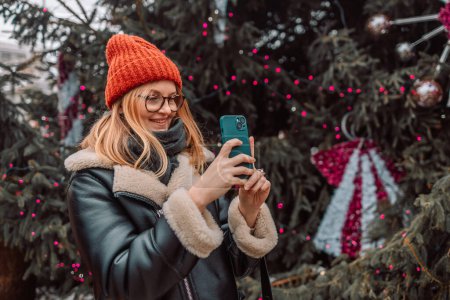 Foto de Hermosa joven feliz toma una foto de un mercado europeo de Navidad con un teléfono inteligente en la noche de invierno. La chica disfruta de la temporada de vacaciones de invierno, visita el mercado de Navidad al aire libre, toma - Imagen libre de derechos