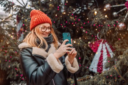 Foto de Mujer rubia feliz toma una foto de un mercado europeo de Navidad con un teléfono inteligente. La chica disfruta de la temporada de vacaciones de invierno, visita el mercado de Navidad al aire libre, toma fotos en un teléfono móvil. - Imagen libre de derechos