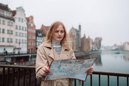 Téléchargez les photos : Attrayant jeune touriste féminine explore la nouvelle ville. Fille blonde tenant une carte sur papier sur la rue de la ville à Gdansk. Voyager en Europe en automne. - en image libre de droit